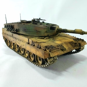 Puede incluir: Maqueta detallada de un tanque militar, con un patrón de camuflaje verde, marrón y amarillo. El tanque tiene un cañón largo, múltiples ruedas y detalles intrincados en el exterior, mostrando un diseño realista.