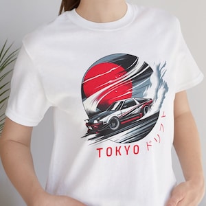 Puede incluir: Camiseta blanca con un estampado gráfico de un sol rojo, un disco negro y blanco y un coche blanco con detalles rojos conduciendo por un camino negro y blanco. El texto "TOKYO ドリフト" está impreso debajo del gráfico.