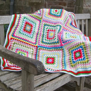 May include: A colourful crocheted blanket with a granny square pattern in shades of red, green, blue, yellow, and white. The blanket is draped over a wooden bench.