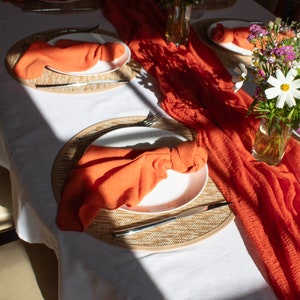 May include: A white tablecloth with orange napkins folded into a decorative shape on white plates. The plates are on woven placemats. A runner of orange fabric is draped across the table. Two bouquets of flowers are in glass vases on the table.