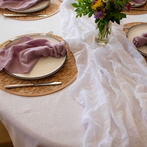 Peut inclure: Un chemin de table blanc à texture plissée, posé sur une nappe blanche. Deux couverts sont visibles, chacun avec un dessous-de-plat en osier, une assiette blanche, une serviette violette et des couverts. Un bouquet de fleurs sauvages dans un vase en verre est placé au centre de la table.