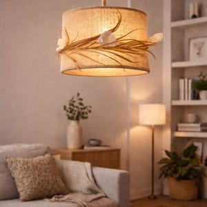 Puede incluir: Una lámpara colgante de tambor beige con una pantalla de tejido natural, adornada con hojas secas y detalles florales blancos. La luz cuelga de un cordón de cuerda, iluminando un salón.