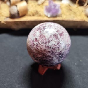 Peut inclure: Une sphère de pierre précieuse polie, avec un motif moucheté de violet et de blanc. La boule de cristal repose sur un petit support en bois brun rougeâtre, sur fond sombre. La sphère est un objet décoratif.