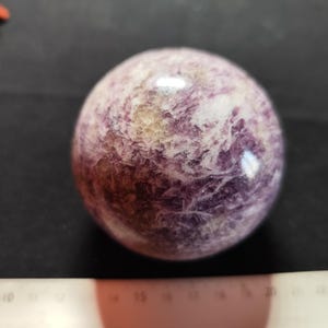 Peut inclure: Une pierre précieuse sphérique et polie avec un aspect marbré. La pierre présente un mélange de teintes violettes, blanches et beiges, créant un motif unique. La surface réfléchit la lumière, soulignant la texture lisse. Un objet décoratif.