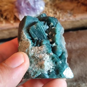May include: A raw mineral specimen featuring deep teal-colored crystalline structures clustered together. The cluster displays sharp, angular facets with a glossy finish, contrasted by a rough, light-colored, textured matrix base at the side.
