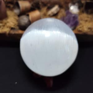 Peut inclure: Une sphère de sélénite blanche avec une surface lisse et polie. La sphère est posée sur un support en bois.