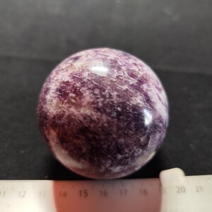 Peut inclure: Une sphère polie de lépidolite, un minéral violet et blanc. La sphère est lisse et réfléchissante, avec un aspect marbré. La couleur est un mélange de violet foncé et de lavande pâle. Une règle est au premier plan, avec des mesures en centimètres.