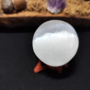 Peut inclure: Une sphère de sélénite blanche, posée sur un petit support en bois brun rougeâtre. La sphère a une surface lisse et polie avec une texture naturelle subtile. Le fond est sombre, mettant en valeur la luminosité de la sphère.