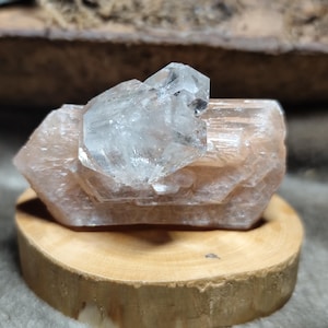 Op de afbeelding: Een heldere kwartskristalgroep ligt bovenop een grotere, perzikkleurige kristalformatie. De kristallen worden gepresenteerd op een kleine, ronde houten basis, die hun natuurlijke schoonheid en geologische structuur benadrukt.