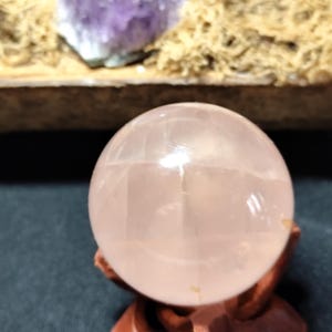 Pode incluir: Uma bola de cristal rosa pálido translúcida, repousando sobre um pequeno suporte de madeira castanha. A esfera é lisa e parece ser feita de quartzo rosa. No fundo, há uma pitada de cristal roxo e musgo seco.