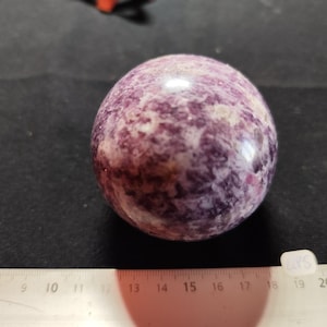 Peut inclure: Une pierre précieuse sphérique polie, aux tons violets et blancs, posée sur une surface sombre. La surface de la pierre présente un aspect marbré. Une règle est placée en dessous.