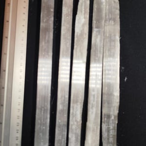 Peut inclure: Cinq baguettes de sélénite, chacune mesurant environ 15 cm de long, sont disposées côte à côte sur une surface noire. Les baguettes sont blanches et translucides, avec une texture légèrement rugueuse. Une règle est visible en arrière-plan pour l'échelle.