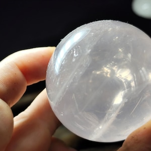May include: A clear, spherical rose quartz crystal ball. The polished sphere has internal veining and is held in a hand, with a dark background. The crystal ball is a pale pink color and reflects light.