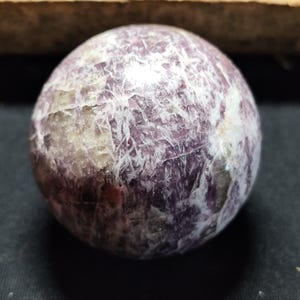 Peut inclure: Une pierre précieuse sphérique et polie. La pierre présente un motif moucheté de couleurs violet, blanc et beige, créant une surface texturée. Il s'agit probablement d'un objet décoratif ou de collection.
