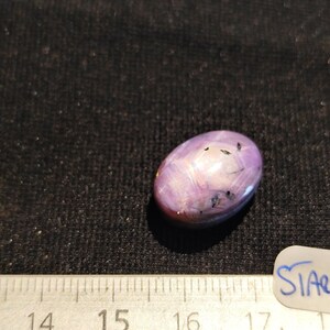 Peut inclure: Pierre précieuse ovale et polie, de couleur rose-violet, avec des inclusions sombres. La pierre est posée sur un fond sombre, avec une règle et une étiquette portant l'inscription "SIARC7".
