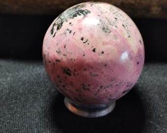 May include: A pink and black sphere made of polished stone. The sphere has a smooth, glossy finish and is speckled with black flecks.