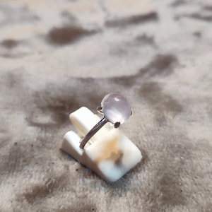 May include: A silver ring with a light pink oval-shaped gemstone. The ring is on a white display stand.