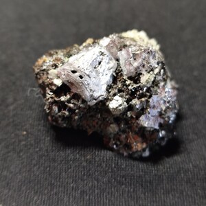 May include: A close-up of a small, rough, dark gray rock with silver-colored crystals embedded in it.