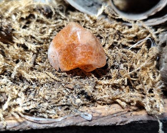 May include: A translucent, orange gemstone with sparkling inclusions sits on dried, light brown moss. The stone has a rounded, irregular shape, and the moss provides a textured, natural backdrop. The image is a close-up.