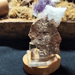 Peut inclure: Une petite roche brune rugueuse avec un minéral blanc cristallin qui lui est attaché. La roche est exposée sur un support en bois avec une base en acrylique transparente.