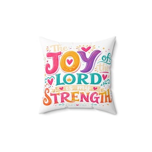 Pode incluir: Uma almofada quadrada branca com texto gráfico colorido que diz The JOY of the LORD is my STRENGTH, com um pequeno ícone de coroa e várias formas de coração em tons de rosa, laranja, roxo, azul-petróleo e amarelo na parte frontal.