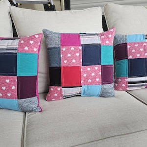May include: Three decorative throw pillows with a patchwork design. The pillows are made from different fabrics in shades of pink, blue, white, and black. The pillows have a white heart pattern on the pink fabric.