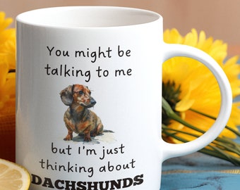 May include: White ceramic coffee mug with a brown and black dachshund illustration. The mug has the text "You might be talking to me but I'm just thinking about DACHSHUNDS" printed on it.
