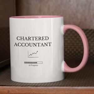 May include: White ceramic mug with a pink rim and handle. The mug has a black graphic that says "CHARTERED ACCOUNTANT" with a line graph and a progress bar that says "In Progress".