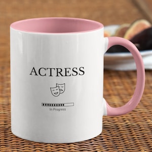 Op de afbeelding: Witte keramische mok met een roze rand en handvat. De mok heeft een zwarte afbeelding met de tekst "ACTRESS" met twee theatermaskers en een voortgangsbalk met de tekst "In Progress".