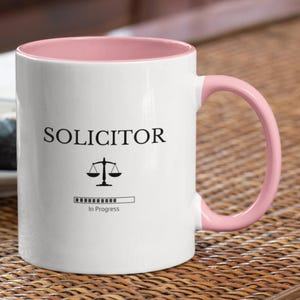 May include: White ceramic mug with a pink rim and handle. The mug has a black graphic of a scale and the text "SOLICITOR" with a progress bar that reads "In Progress".
