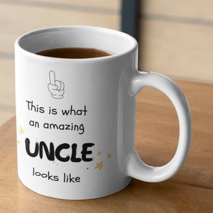 Puede incluir: Taza de cerámica blanca con asa negra, llena de líquido oscuro. La taza presenta el texto "This is what an amazing UNCLE looks like" con un gráfico de dedo señalando y estrellas doradas.