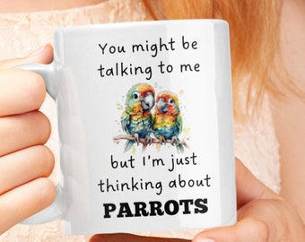 May include: White ceramic mug with a colorful illustration of two parrots on a branch. The mug has the text "You might be talking to me but I'm just thinking about PARROTS" printed on it.