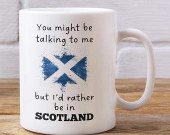 Może przedstawiać: Biały ceramiczny kubek z zakrzywionym uchwytem. Kubek zawiera napis "You might be talking to me but I'd rather be in SCOTLAND" z niebiesko-białym wzorem flagi szkockiej. Kubek stoi na drewnianej powierzchni.