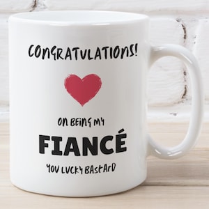 May include: White ceramic mug with black text that reads "Congratulations! On being my fiancé You lucky bastard" and a red heart.
