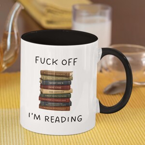 May include: White coffee mug with a black rim and handle. The mug has the text "FUCK OFF I'M READING" printed on it. A stack of books with titles like "Wuthering Heights", "Pride and Prejudice", "Moby Dick", "Jane Eyre", "The Odyssey", "Great Expectations", "The Picture of Dorian Gray", "Adventures of Huckleberry Finn", and "Crime and Punishment" are printed on the mug.
