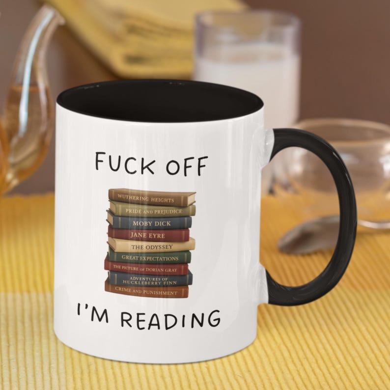 May include: White coffee mug with a black rim and handle. The mug has the text "FUCK OFF I'M READING" printed on it. A stack of books with titles like "Wuthering Heights", "Pride and Prejudice", "Moby Dick", "Jane Eyre", "The Odyssey", "Great Expectations", "The Picture of Dorian Gray", "Adventures of Huckleberry Finn", and "Crime and Punishment" are printed on the mug.