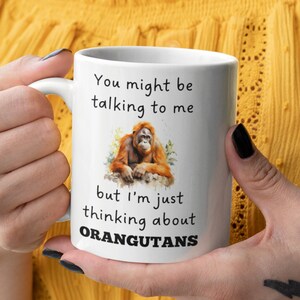 May include: White ceramic mug with a black and white graphic of an orangutan. The text on the mug reads "You might be talking to me but I'm just thinking about ORANGUTANS."
