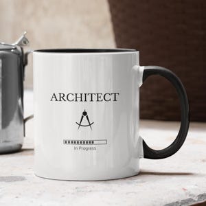 Peut inclure: Mug en céramique blanche avec une anse et un bord noirs. Le mug porte le mot "ARCHITECTE" en noir, ainsi qu'un symbole de compas et un graphique de barre de chargement "En cours". Le mug est conçu pour les architectes.