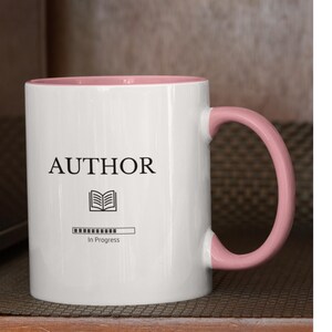 May include: White ceramic mug with a pink rim and handle. The mug has the text "AUTHOR" printed on it, along with a book icon and a progress bar that reads "In Progress".