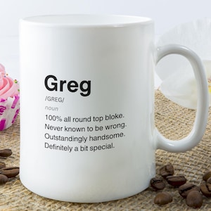 Może przedstawiać: Biały ceramiczny kubek z napisem "Greg" i definicją wydrukowaną na czarno. Definicja zawiera zwroty takie jak "100% all round top bloke" i "Outstandingly handsome". Babeczka z różowym lukrem i ziarnami kawy w tle.