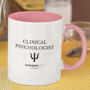 Può includere: Una tazza in ceramica bianca con bordo e manico rosa. La tazza ha il testo "Clinical Psychologist" con un simbolo di psicologia e una barra di avanzamento che recita "In Progress".