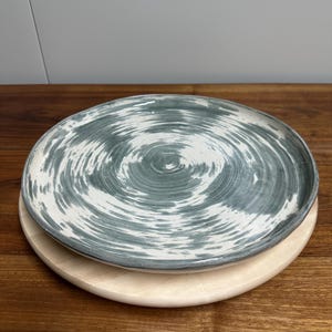Peut inclure: Une assiette en céramique faite à la main avec un motif en spirale gris et blanc, posée sur une planche de service ronde en bois. L'assiette a un bord légèrement surélevé et une finition mate, idéale pour servir des aliments.