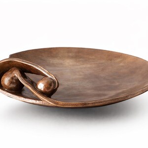 Op de afbeelding: Een bronzen decoratieve kom met een ovale vorm. De kom heeft een uniek ontwerp met twee bolvormige accenten genesteld in een gebogen, bladachtig element. De algehele esthetiek is modern en artistiek.