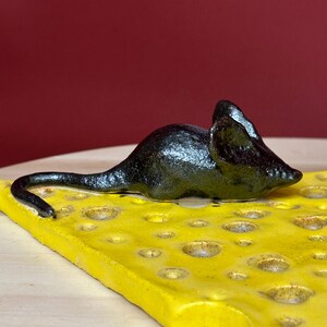 Op de afbeelding: Een keramische sculptuur van een zwarte muis die rust op een gele kaaswig. De kaas heeft verschillende gaten en staat tegen een rode achtergrond. De muis heeft een lange staart en puntige oren.