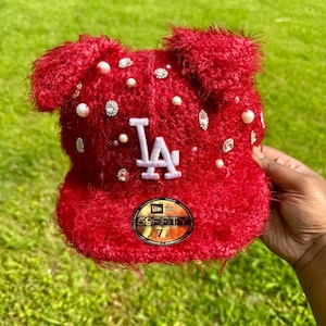 May include: A red, fuzzy baseball cap with the letters "LA" in white. The cap is embellished with pearls and rhinestones. The cap has a "59FIFTY" sticker on the brim.