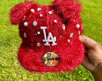 May include: A red, fuzzy baseball cap with the letters "LA" in white. The cap is embellished with pearls and rhinestones. The cap has a "59FIFTY" sticker on the brim.