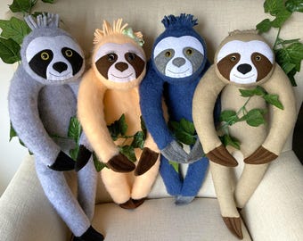 May include: Four plush sloth toys in different colours: grey, peach, blue, and tan. The sloths are all in a sitting position with their arms outstretched.