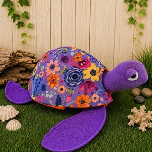 May include: A plush turtle toy featuring a purple felt head and flippers with a quilted texture. The shell is covered in a vibrant floral fabric pattern displaying orange, yellow, pink, and blue flowers on a purple background with an orange trim.