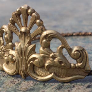May include: A gold-colored, ornate tiara with a detailed, vintage design. The tiara features a central, fan-shaped element and swirling, floral motifs. The band is a twisted, bronze-colored metal. The piece is likely for a special occasion.