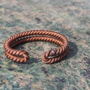 Op de afbeelding: Een koperkleurige ring met een gedraaid touwontwerp. De ring is aan de voorkant open en heeft een kleine, ronde versiering. De ring ligt op een gevlekte groen-grijze ondergrond.